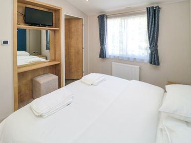 A bedroom with a bed covered in white linens two folded towels a window with curtains a wooden door a wall-mounted television and a cushioned stool at 28 Finch in Cayton Bay