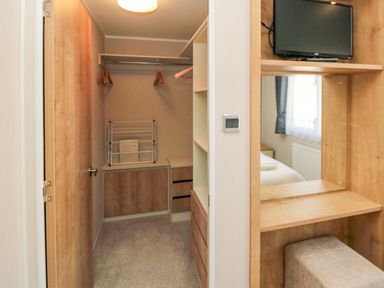 A walk-in closet with hangers a drying rack and wooden cabinets next to a vanity area with a mirror a stool and a mounted television at 28 Finch in Cayton Bay