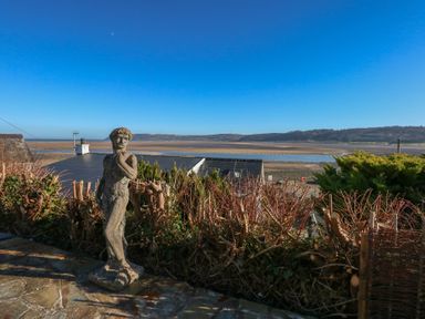 A garden patio with a stone statue overlooking rooftops hills and a body of water at Swn Y Wylan in Red Wharf Bay near Benllech