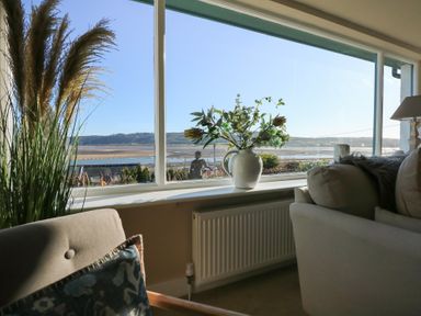 A living room with plants and flowers near a large window overlooking a riverside landscape at Swn Y Wylan Red Wharf Bay near Benllech