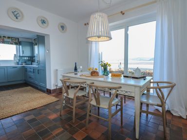 A dining area with a table set with food and drinks and chairs next to a window and an adjoining kitchen at Swn Y Wylan Red Wharf Bay near Benllech