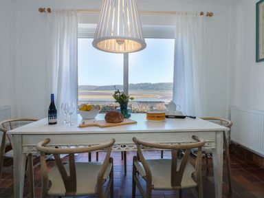 A dining room with a table set with bread a cake wine glasses and a vase of flowers by a large window at Swn Y Wylan Red Wharf Bay near Benllech