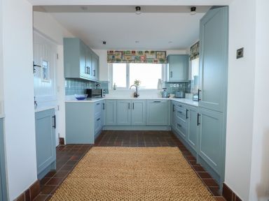 A kitchen with blue cabinets a window above the sink a brown tiled floor and a woven rug on the floor at Swn Y Wylan in Red Wharf Bay near Benllech