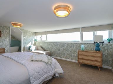 A bedroom with a bed a chaise lounge a wooden dresser with a lamp and a bathroom visible through an open door at Swn Y Wylan Red Wharf Bay near Benllech