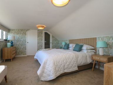 A bedroom with a double bed with white bedding and turquoise pillows two wooden bedside tables with turquoise lamps a wooden dresser with a lamp and window on one side at Swn Y Wylan Red Wharf Bay near Benllech