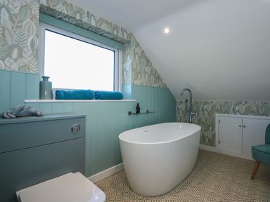 A bathroom with a white freestanding bathtub blue cabinet blue towels and leaf patterned wallpaper at Swn Y Wylan Red Wharf Bay near Benllech
