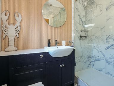 A bathroom with a round mirror a sink on a cabinet a shower with glass door and marble wall tiles and a lobster decoration on the wall at Swn Y Wylan in Red Wharf Bay near Benllech
