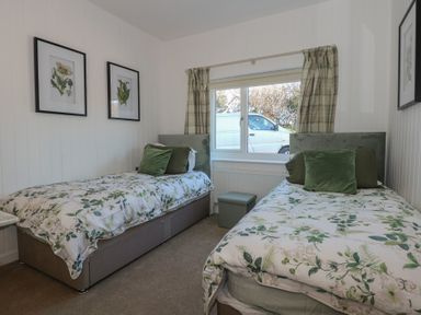 A bedroom with two single beds with green and white floral bedding and green pillows and a window with checkered curtains at Swn Y Wylan Red Wharf Bay near Benllech