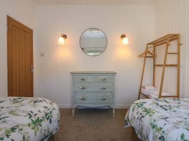 A bedroom with two beds with leaf patterned bedding a wooden chest of drawers a round wall mirror a wooden door and a wooden clothes rack with towels and hangers at Swn Y Wylan Red Wharf Bay near Benllech
