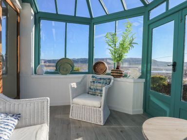 A sunroom with white wicker chairs and green framed windows overlooking a landscape at Swn Y Wylan in Red Wharf Bay near Benllech