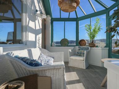 A sunroom with white wicker furniture and blue cushions and plants in pots at Swn Y Wylan Red Wharf Bay near Benllech