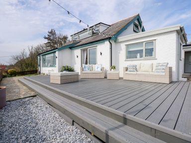 A raised outdoor deck with wicker sofas and cushions outside a white house at Swn Y Wylan Red Wharf Bay near Benllech