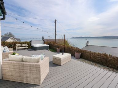 An outdoor deck with wicker furniture string lights a hot tub and a view of the sea at Swn Y Wylan in Red Wharf Bay near Benllech