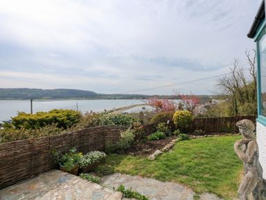 A garden area with plants a stone path and a statue overlooking a body of water with hills in the background at Swn Y Wylan Red Wharf Bay near Benllech