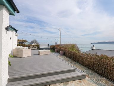 An outdoor deck with wicker furniture and string lights overlooking the sea at Swn Y Wylan Red Wharf Bay near Benllech