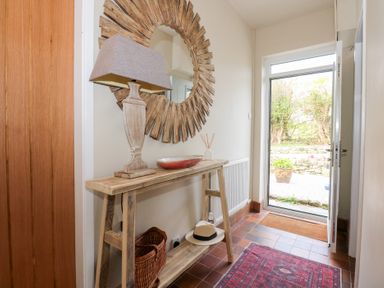 An entryway with a wooden table holding a lamp a bowl and reed diffuser below the table a basket and a hat with a round wooden mirror on the wall and an open door showing an outside area at Swn Y Wylan Red Wharf Bay near Benllech