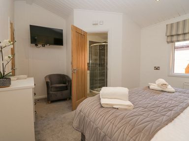 A bedroom with a gray quilted bedspread, towels on the bed, a gray armchair, a wall-mounted TV, a dresser with a plant, and an open door leading to a bathroom with a shower at Lakeland View Ambleside 86 White Cross Bay near Windermere