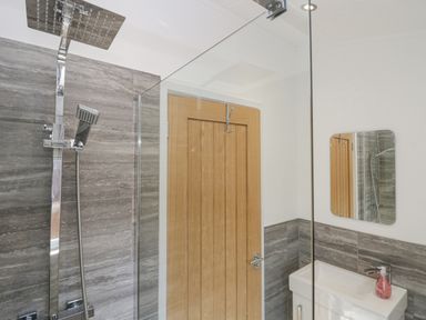 A bathroom with a shower area having a rainfall showerhead a wooden door a small rectangular mirror above a white sink with a pink soap dispenser at Lakeland View Ambleside 86 White Cross Bay near Windermere