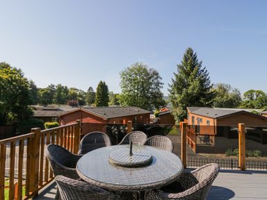 An outdoor patio with a round glass table and wicker chairs overlooking wooden cabins and trees at Lakeland View Ambleside 86 White Cross Bay near Windermere