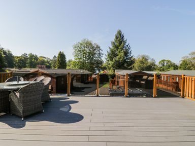 An outdoor deck with a glass table and four wicker chairs overlooking wooden cabins and trees at Lakeland View Ambleside 86 White Cross Bay near Windermere