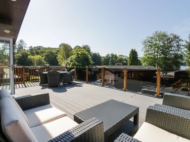An outdoor deck with wicker seating and a dining table overlooking trees and wooden buildings at Lakeland View Ambleside 86 White Cross Bay near Windermere