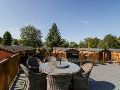 An outdoor deck with a round glass table and six wicker chairs overlooking wooden cabins and trees at Lakeland View in Ambleside 86 White Cross Bay near Windermere
