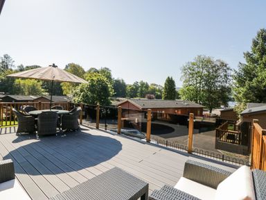 A balcony with outdoor furniture and umbrella overlooking wooden cabins and trees at Lakeland View Ambleside 86 White Cross Bay near Windermere