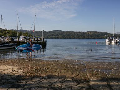 A man carrying a paddleboard in shallow water near boats docked at a pier by a lake with wooded hills in the background at Lakeland View Ambleside 86 White Cross Bay near Windermere