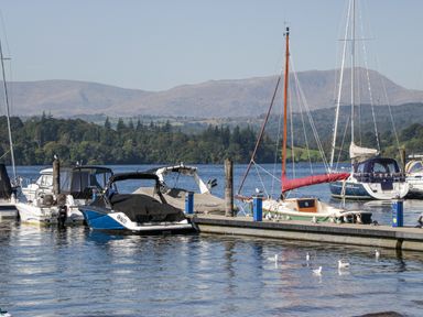 A marina with several boats docked and birds on the water and pier with hills in the background at Lakeland View in Ambleside 86 White Cross Bay near Windermere