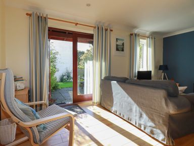 A living room with a gray sofa, a wooden armchair with cushions, striped curtains, a floor lamp, and glass doors opening to a garden at Coombe Cottage in Falmouth