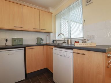 A kitchen with wooden cabinets a white dishwasher a small fridge a green toaster and kettle and a window with blinds at Coombe Cottage in Falmouth