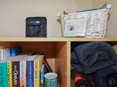 A wooden shelf with books and folded blankets inside and a black bag and white basket on top at Coombe Cottage Falmouth