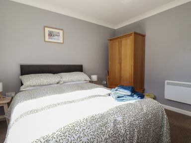 A bedroom with a double bed pillow and blanket with leaf print a wooden wardrobe two bedside tables with lamps and a framed picture on the wall at Coombe Cottage in Falmouth