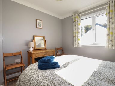 A bedroom with a bed covered in patterned bedding and blue towels on top wooden chairs a wooden dresser with a mirror and a lamp and a window with curtains at Coombe Cottage in Falmouth