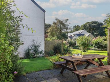 A garden with a wooden picnic table on a paved area beside a white house with greenery at Coombe Cottage in Falmouth
