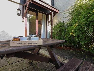 A wooden picnic table with a tray holding cups and jars outside a house with a glass door and greenery at Coombe Cottage in Falmouth
