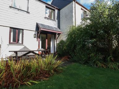 A garden area with green grass and bushes outside a two-story house with white walls and brown windows at Coombe Cottage in Falmouth