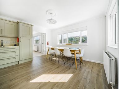 A dining area with a wooden table and white chairs next to windows overlooking hills at Fairwinds in Portreath