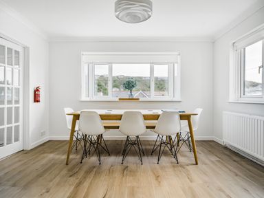 A dining room with a wooden table and six white chairs near windows at Fairwinds in Portreath