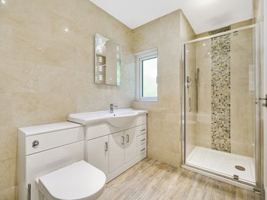 A bathroom with a toilet, sink with storage cabinets, a mirror, a window, and a glass-enclosed shower with mosaic tiles at Fairwinds in Portreath