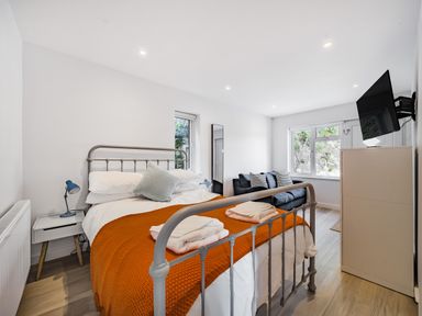 A bedroom with a metal bed frame orange blanket towels bedside table blue lamps sofa wall-mounted tv and window at Fairwinds in Portreath