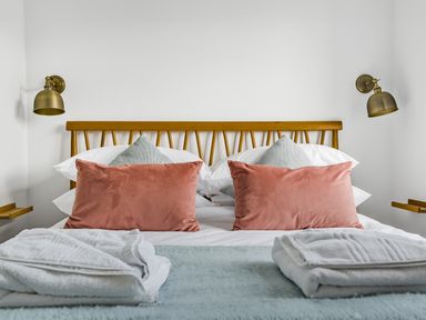 A bed with wooden headboard pink and gray pillows white towels and brass wall lamps at Fairwinds in Portreath