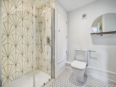 A bathroom with a shower enclosure featuring geometric patterned tiles a toilet with hexagonal floor tiles a round mirror and a white door at Fairwinds in Portreath