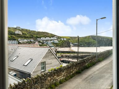 View of houses on a hillside overlooking a beach and sea with a road and stone wall in foreground at Fairwinds in Portreath
