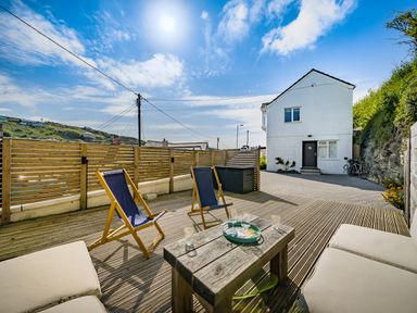 An outdoor wooden deck with two blue deck chairs a wooden table with glasses and a tray a white cushioned bench and a white house in the background at Fairwinds in Portreath