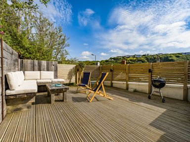 An outdoor deck with a wooden bench with cushions a small table two folding chairs and a barbecue grill at Fairwinds in Portreath