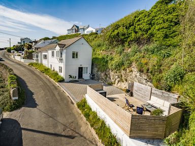 A road beside a white house with a patio and seating area next to a steep hill with green vegetation at Fairwinds in Portreath