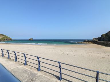 A sandy beach with a blue railing in the foreground and the ocean with a small rock formation at Fairwinds in Portreath