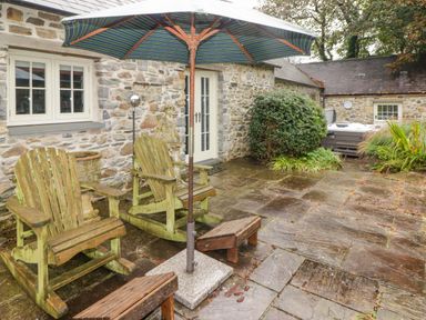 An outdoor patio with two wooden rocking chairs under a patio umbrella stone flooring and stone walls with bushes and a hot tub at St Dogwells Barn near Wolfscastle