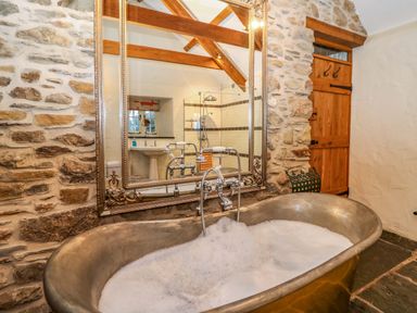 A bathroom with a filled metal bathtub stone walls a large mirror a wooden door and a shower reflected in the mirror at St Dogwells Barn near Wolfscastle